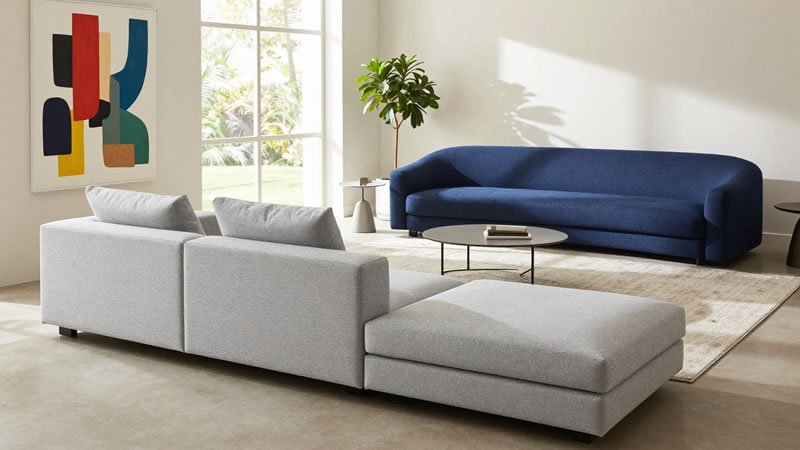 Light gray modular sectional sofa with ottoman and navy blue curved sofa in a spacious modern living room with abstract wall art and indoor plant